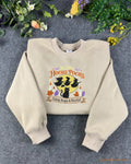 Flat lay of a sand beige Halloween crewneck sweatshirt embroidered with A Little Hocus Pocus, black cat, and flying witches