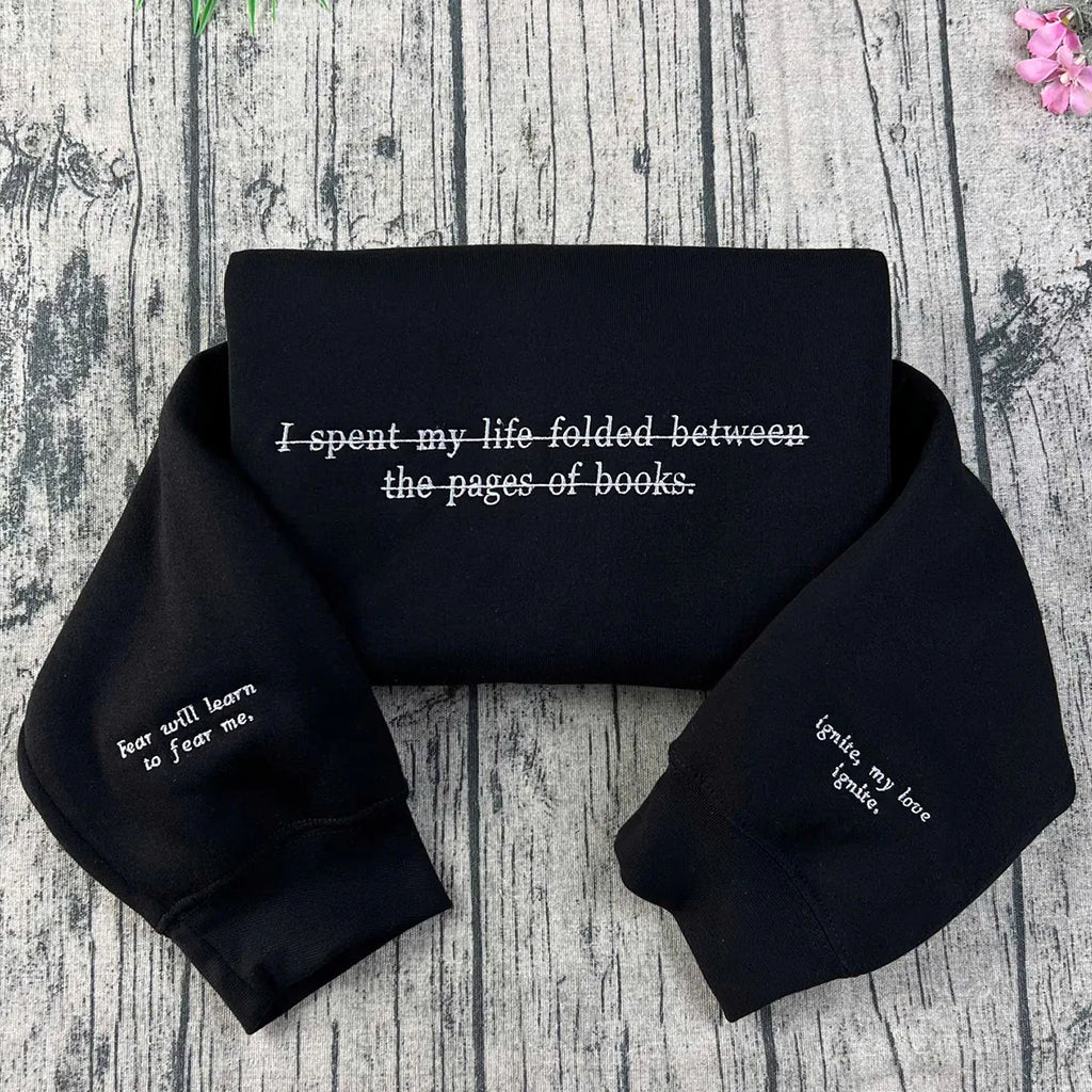 Black crewneck sweatshirt with white embroidered text and quotes from the 'Shatter Me' series, laid flat on a wooden background with a pink flower at the top right corner.