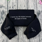 Black crewneck sweatshirt with white embroidered text and quotes from the 'Shatter Me' series, laid flat on a wooden background with a pink flower at the top right corner.