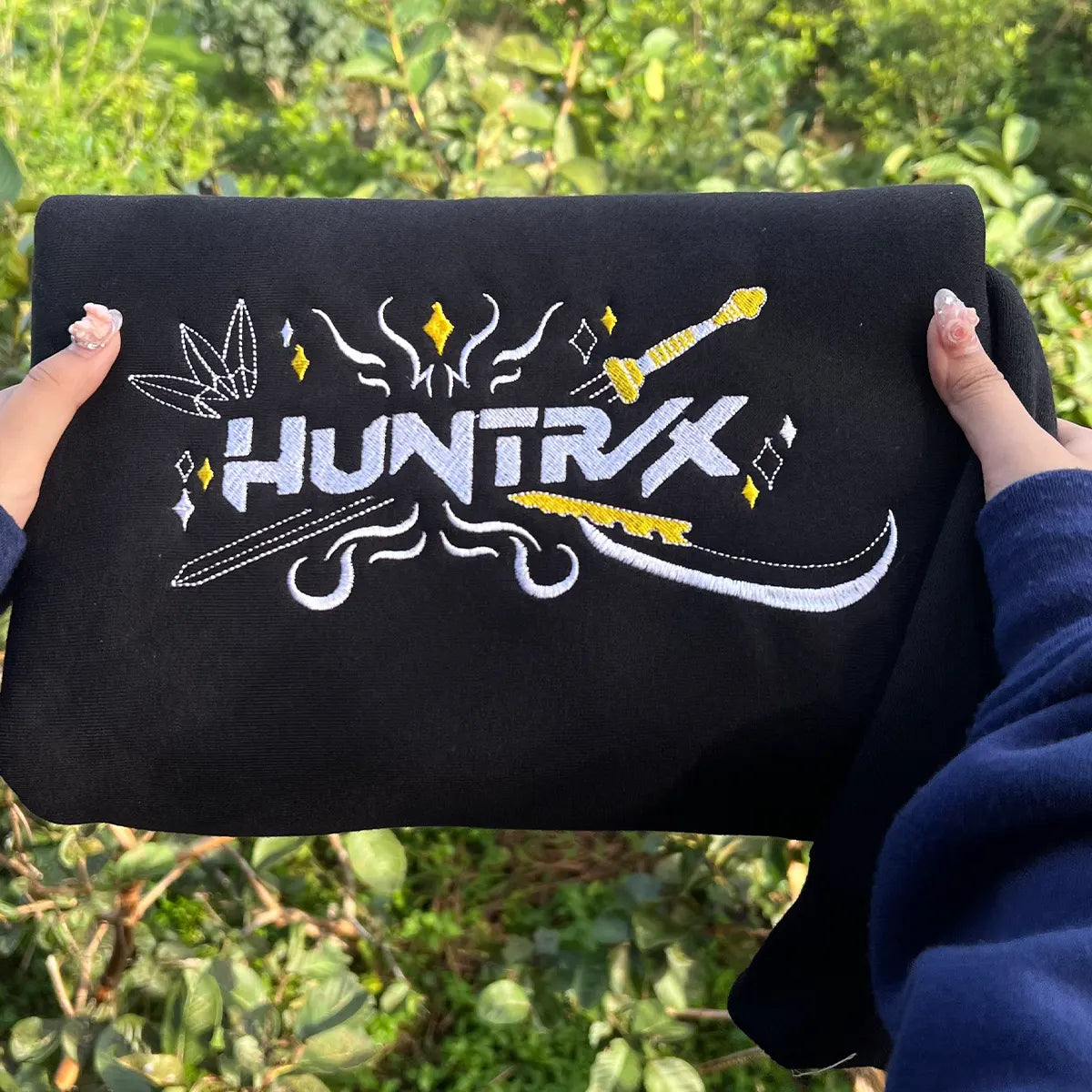 Black pouch held by hands with 'HUNTRX' logo against a green leafy background