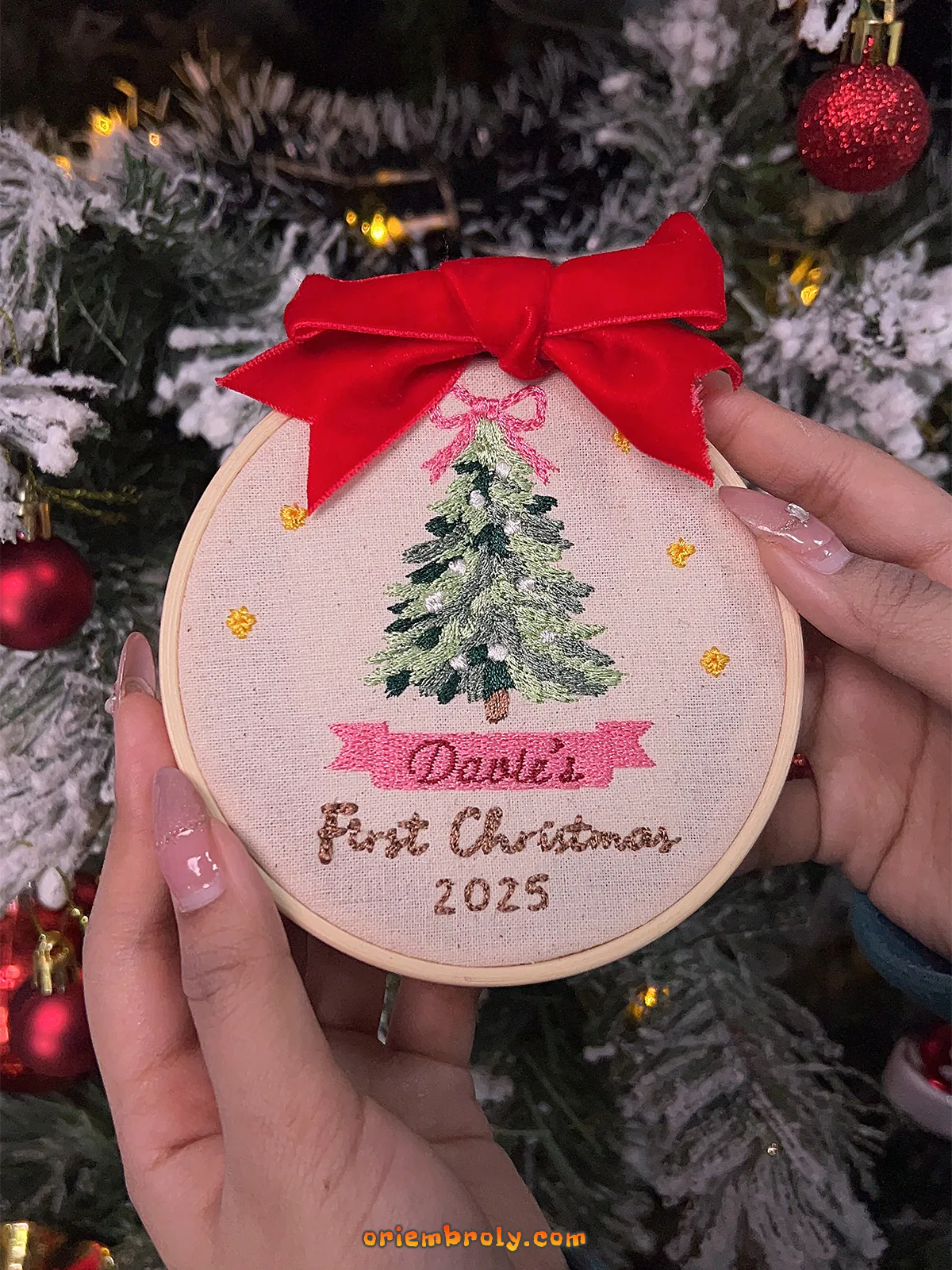 Embroidery hoop with Christmas tree design and text held in front of a decorated tree.