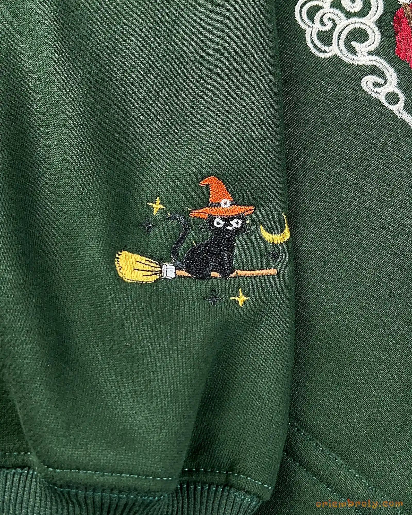 Close-up of custom sleeve embroidery on a green hoodie showing a black cat wearing an orange witch hat riding a broom.