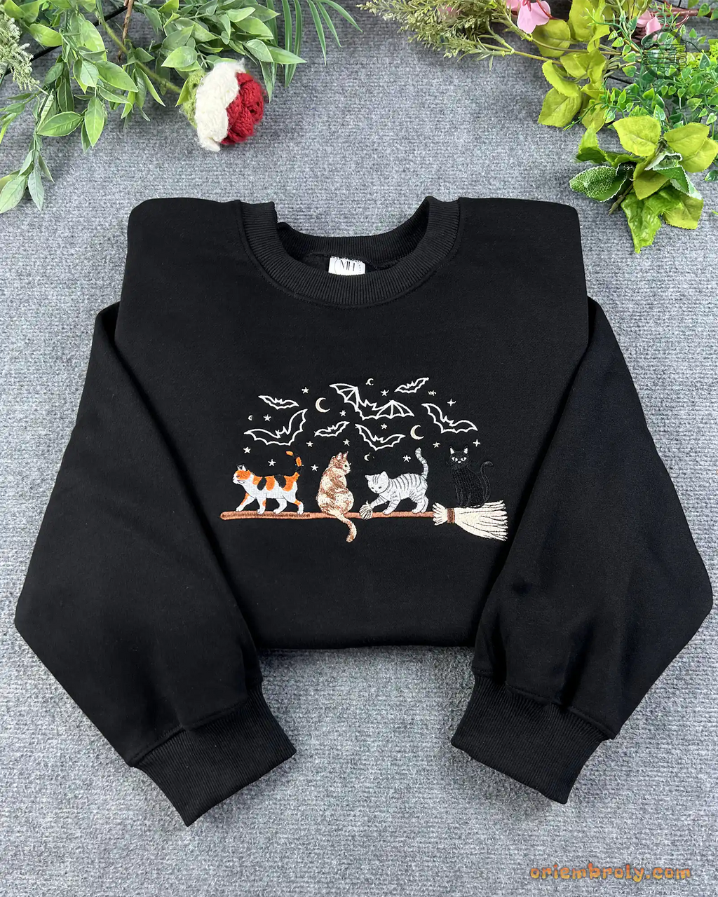 Flat lay of a black Halloween crewneck sweater embroidered with calico, tabby, and black cats on a broomstick
