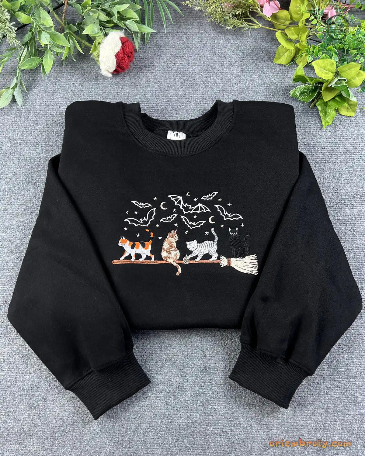 Flat lay of a black Halloween crewneck sweater embroidered with calico, tabby, and black cats on a broomstick
