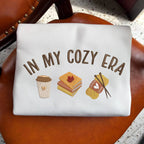 White pouch with 'In My Cozy Era' text and illustrations on a brown surface.
