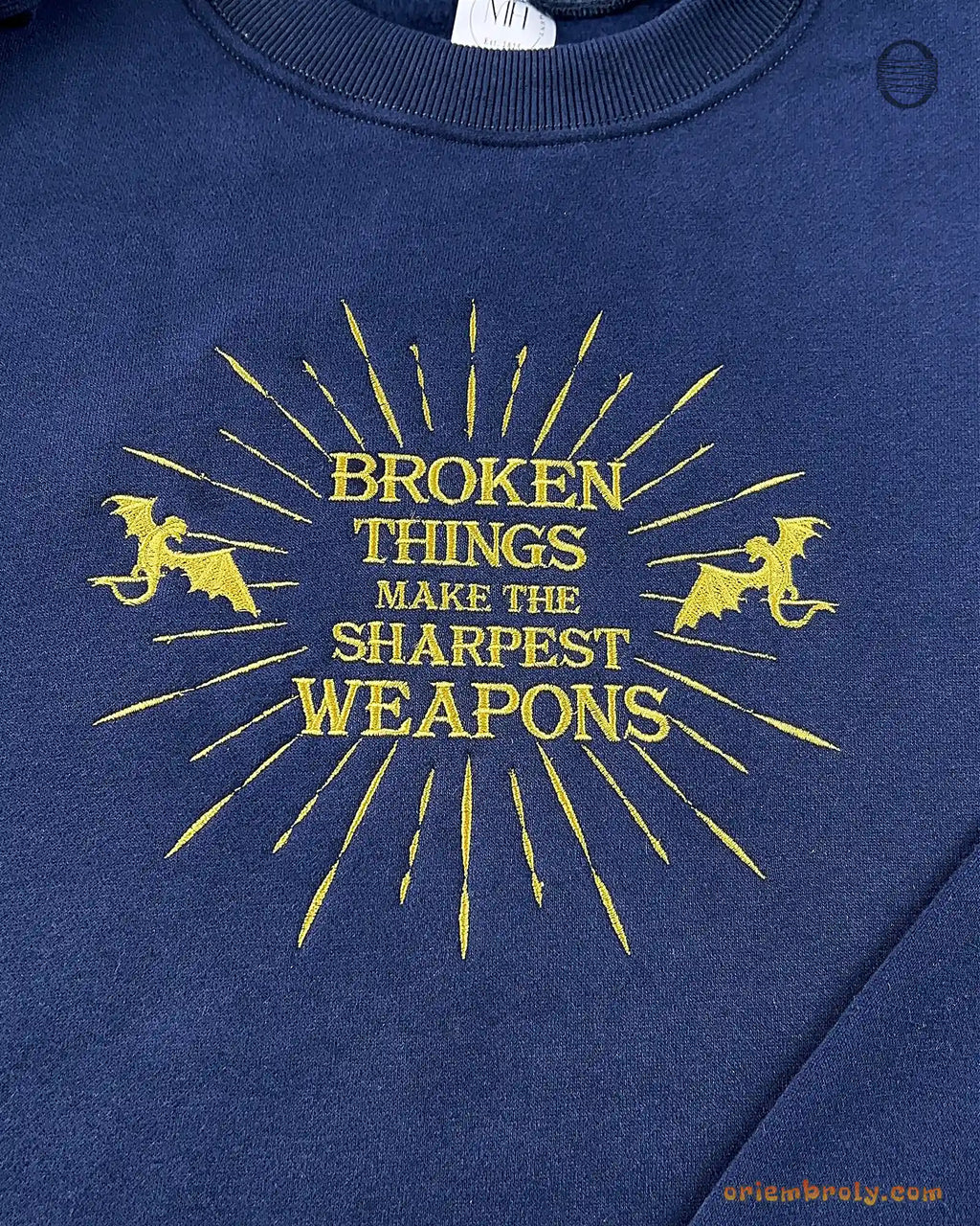 Close-up of gold sunburst burst, pegasus silhouettes, and Broken Things Make The Sharpest Weapons embroidered quote on a navy blue sweater