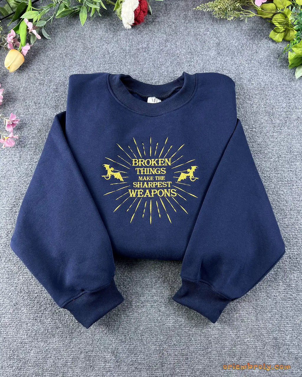 Flat lay of a navy blue crewneck sweatshirt embroidered with gold sunburst pegasus design and Broken Things Make The Sharpest Weapons quote