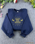 Flat lay of a navy blue crewneck sweatshirt embroidered with gold sunburst pegasus design and Broken Things Make The Sharpest Weapons quote