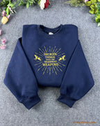 Flat lay of a navy blue crewneck sweatshirt embroidered with gold sunburst pegasus design and Broken Things Make The Sharpest Weapons quote