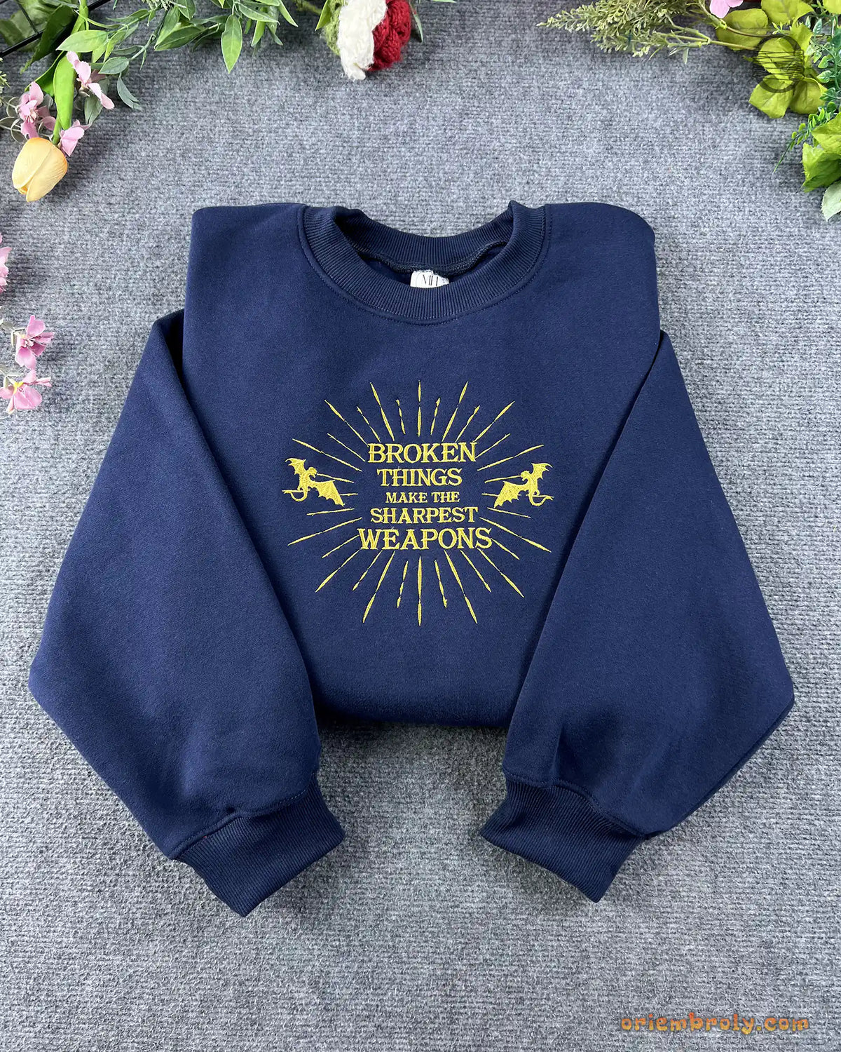 Flat lay of a navy blue crewneck sweatshirt embroidered with gold sunburst pegasus design and Broken Things Make The Sharpest Weapons quote