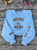 Light blue sweatshirt with 'Chestnut Springs' design on a wooden surface with flowers.