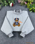 Flat lay of a sport grey crewneck sweatshirt embroidered with a cute teddy bear wearing a navy and orange Chicago football helmet with jersey 34