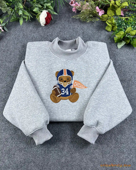 Flat lay of a sport grey crewneck sweatshirt embroidered with a cute teddy bear wearing a navy and orange Chicago football helmet with jersey 34