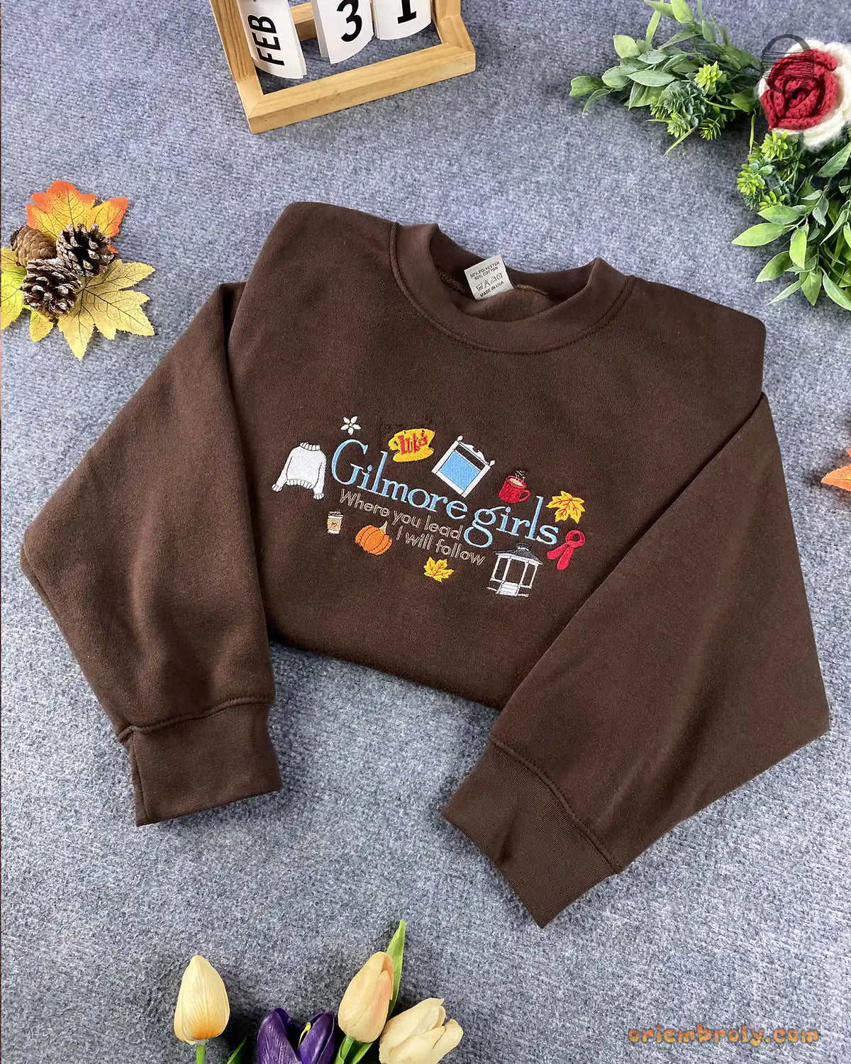 Overview flat lay of a chocolate brown Gilmore Girls embroidered crewneck sweatshirt with fall foliage decor
