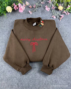 Flat lay of a chocolate brown crewneck sweatshirt embroidered with red Merry Christmas text, a coquette ribbon bow, and Christmas collar icons