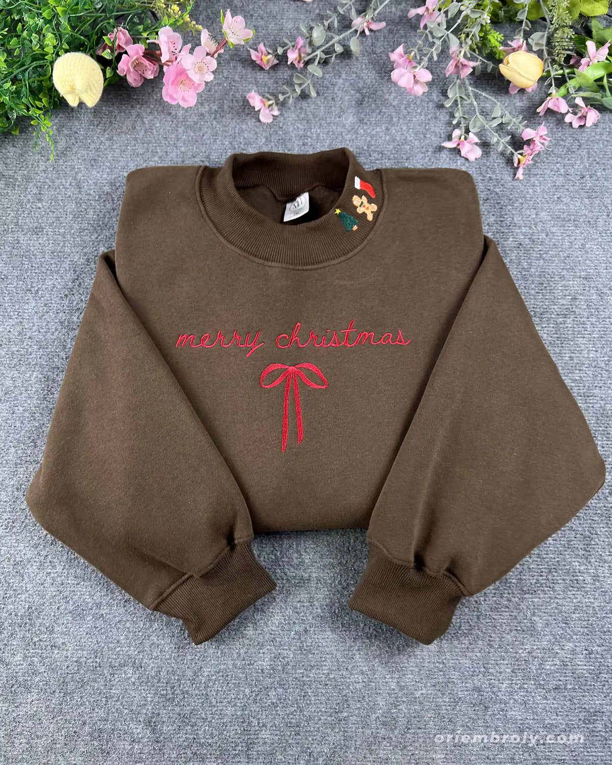 Flat lay of a chocolate brown crewneck sweatshirt embroidered with red Merry Christmas text, a coquette ribbon bow, and Christmas collar icons