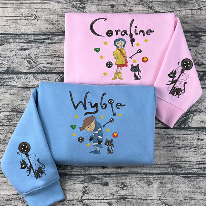 Two hoodies, one pink and one blue, with embroidered characters and text on a wooden surface.