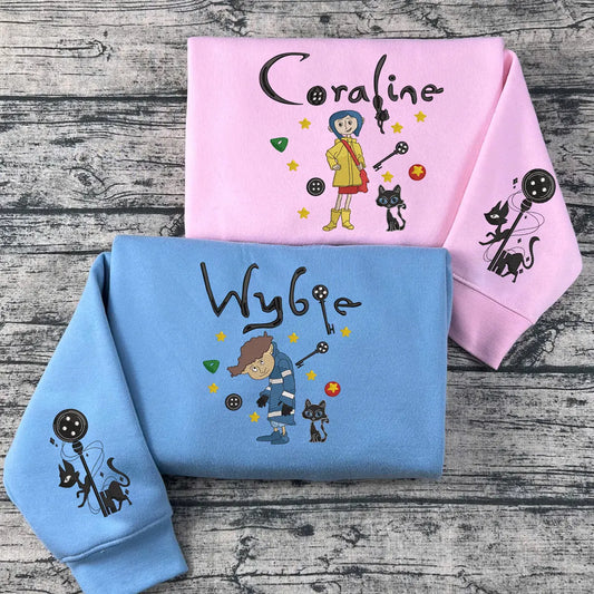 Two hoodies, one pink and one blue, with embroidered characters and text on a wooden surface.