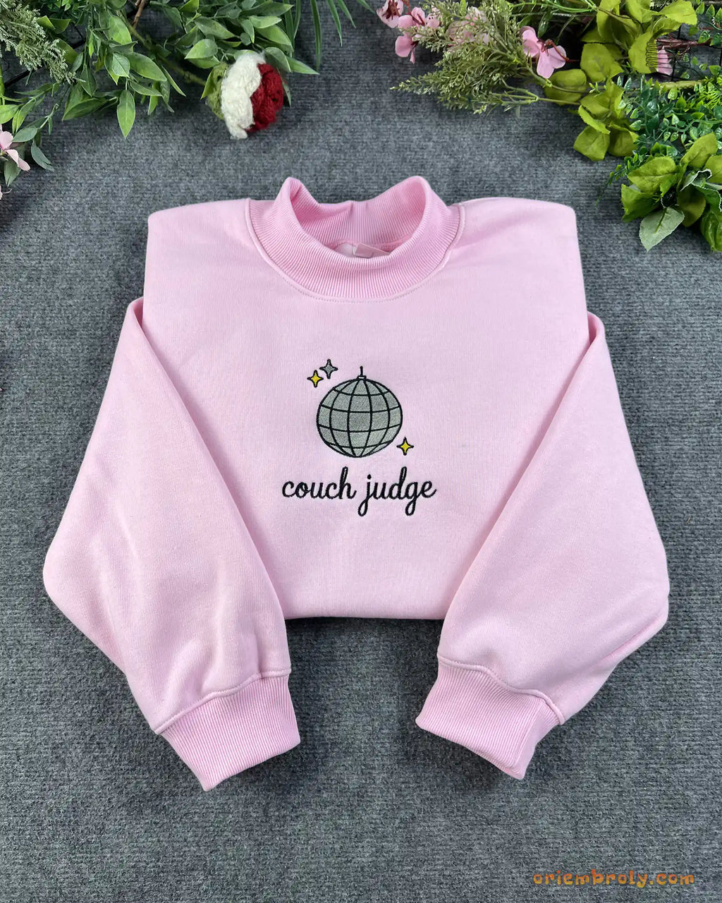 Flat lay of a light pink crewneck sweatshirt embroidered with Couch Judge text and a sparkling disco ball