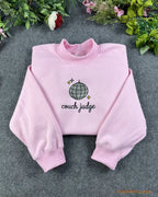 Flat lay of a light pink crewneck sweatshirt embroidered with Couch Judge text and a sparkling disco ball