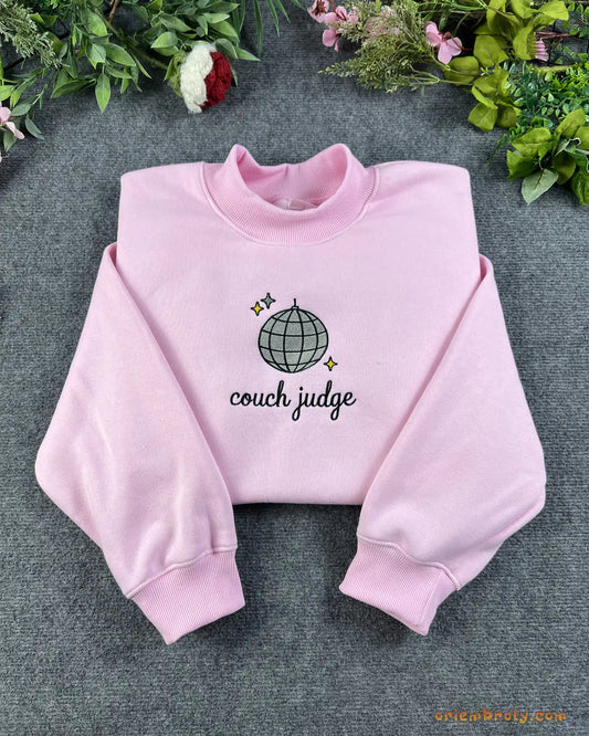 Flat lay of a light pink crewneck sweatshirt embroidered with Couch Judge text and a sparkling disco ball