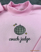 Close-up of silver disco ball with gold sparkles and Couch Judge cursive embroidery on a light pink sweater
