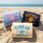 Three beach towels with personalized names and dates on a sandy beach.