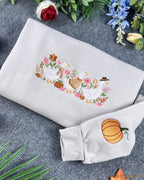Angled view of a light beige crewneck sweatshirt with embroidered ducks and fall floral motifs.
