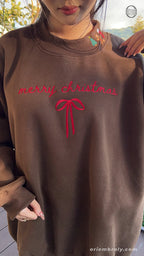 Front view of model wearing a chocolate brown Christmas sweatshirt showing red Merry Christmas script and bow embroidery