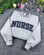 Overview of a sport grey nurse embroidered crewneck sweater featuring varsity block letters and medical stethoscope sleeve embroidery