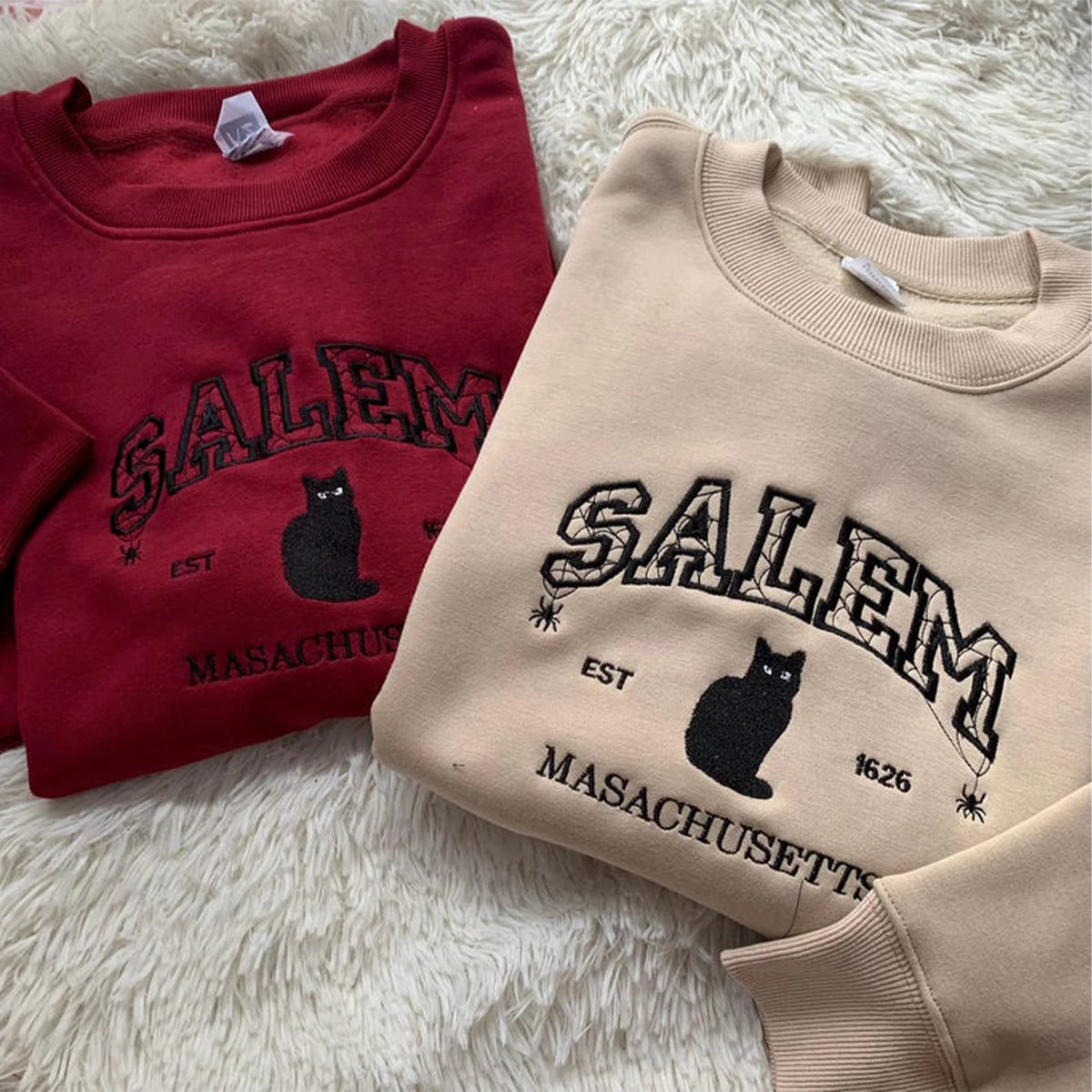 Two sweatshirts with 'Salem, Massachusetts' text and black cat design on a fluffy white surface.