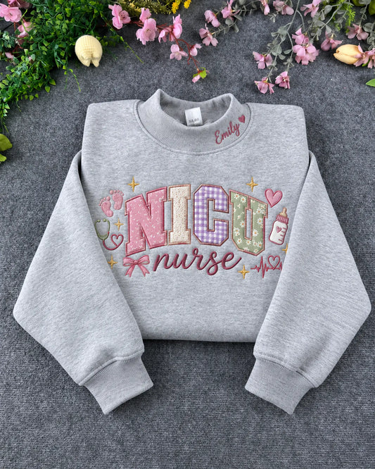 Flat lay of a sport grey crewneck sweatshirt embroidered with pastel NICU nurse pattern, baby footprints, bottle, and custom collar name