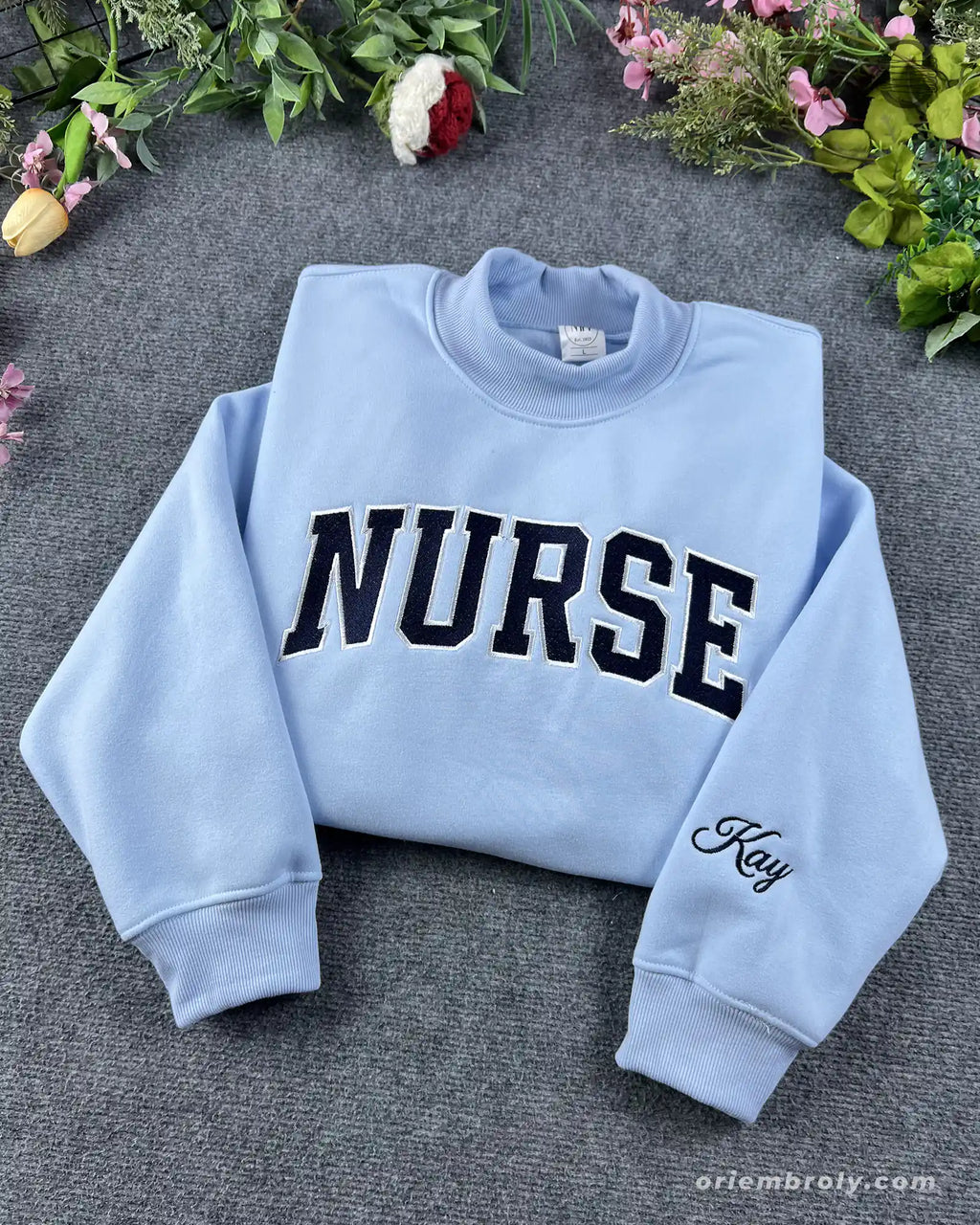 Flat lay of a light blue crewneck sweatshirt with black varsity nurse block lettering and personalized sleeve embroidery