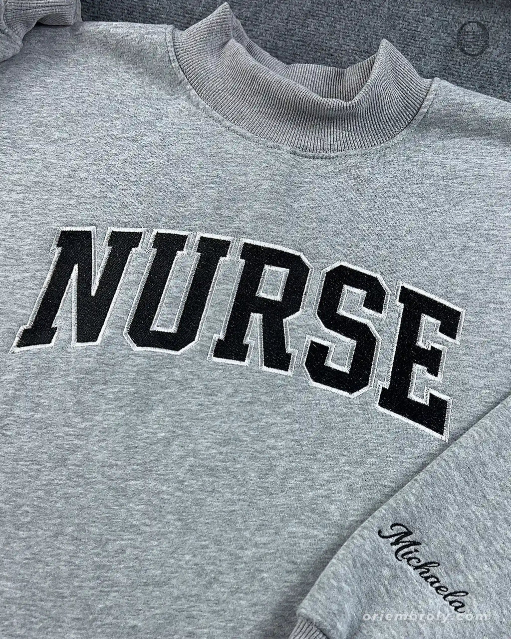 Close-up of black varsity nurse chest embroidery and personalized custom name cursive sleeve embroidery on a grey sweater