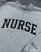 Close-up of black varsity nurse chest embroidery and personalized custom name cursive sleeve embroidery on a grey sweater