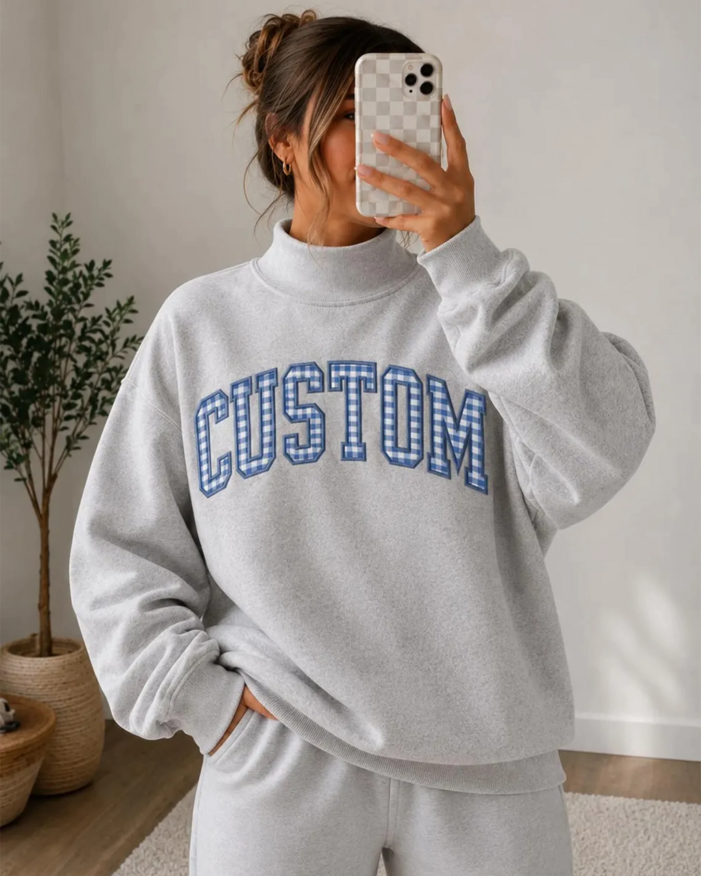 Model taking a selfie wearing a grey mock neck sweatshirt with custom blue plaid embroidered patch letters