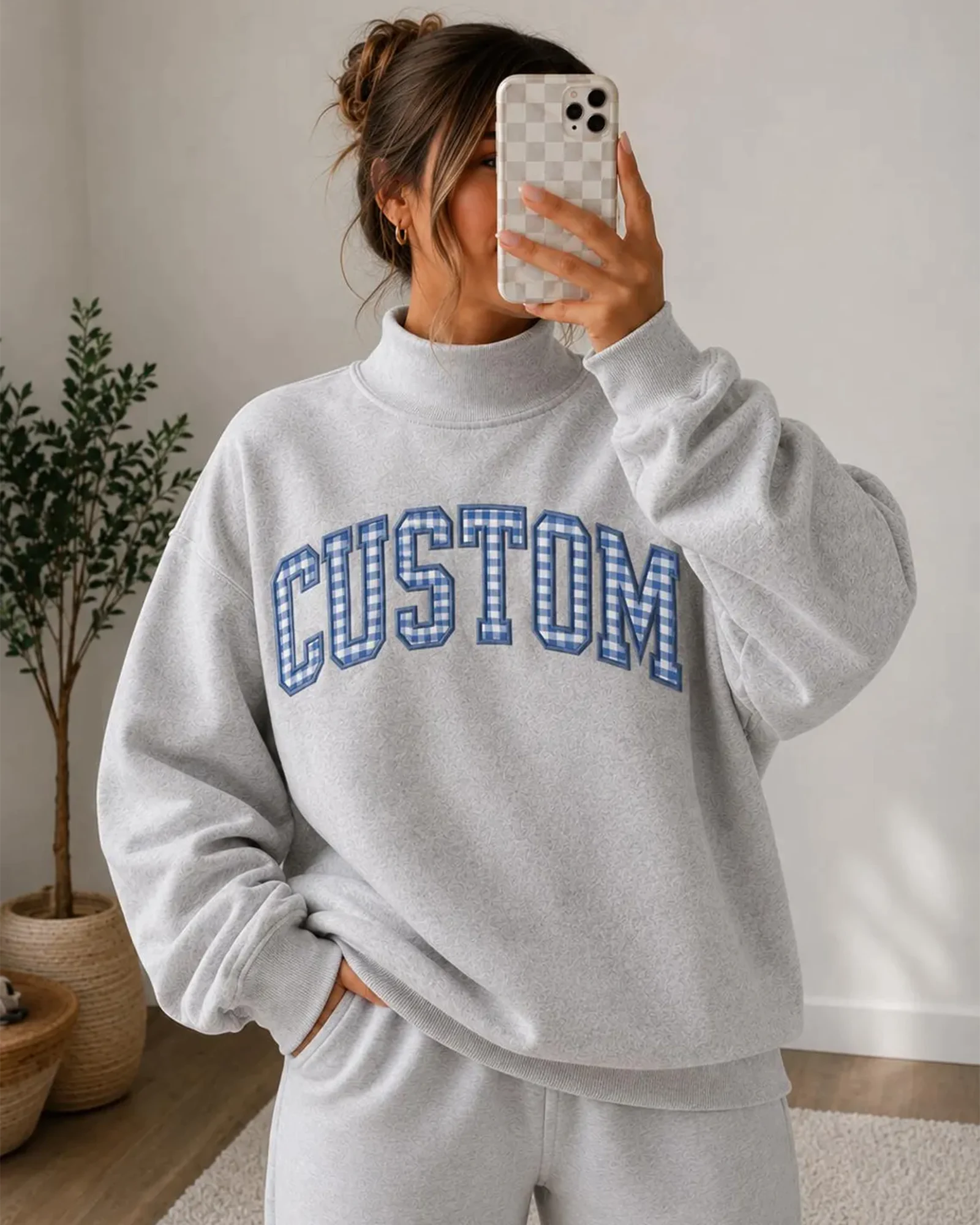 Model taking a selfie wearing a grey mock neck sweatshirt with custom blue plaid embroidered patch letters