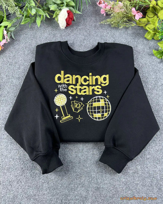 Flat lay of a black crewneck sweatshirt embroidered with gold Dancing with the Stars text, trophy, and mirrorball