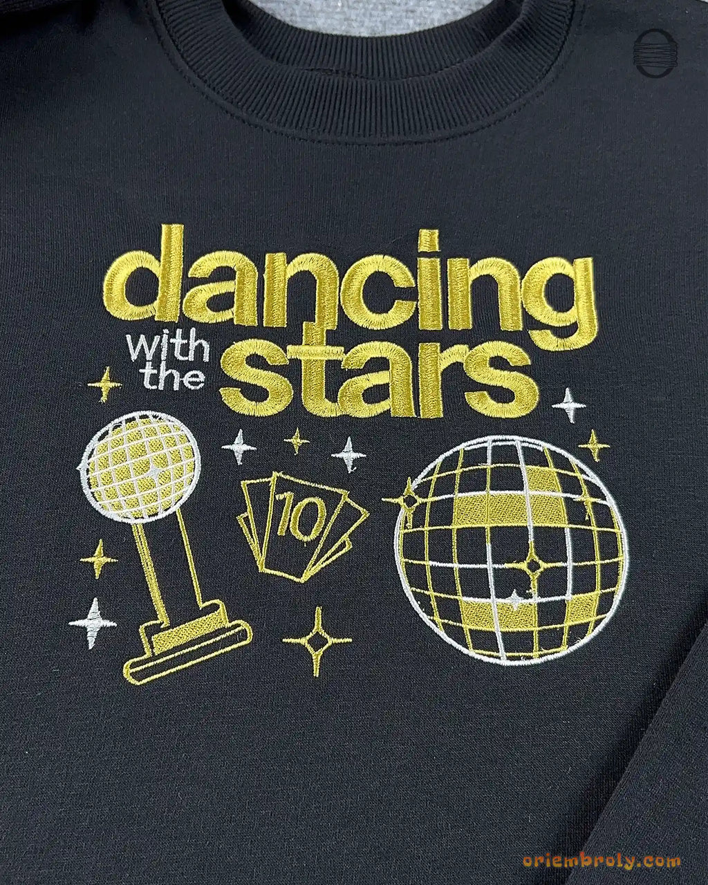 Close-up of gold Dancing with the Stars embroidery featuring mirrorball trophy, dance shoes, and disco ball on a black sweater