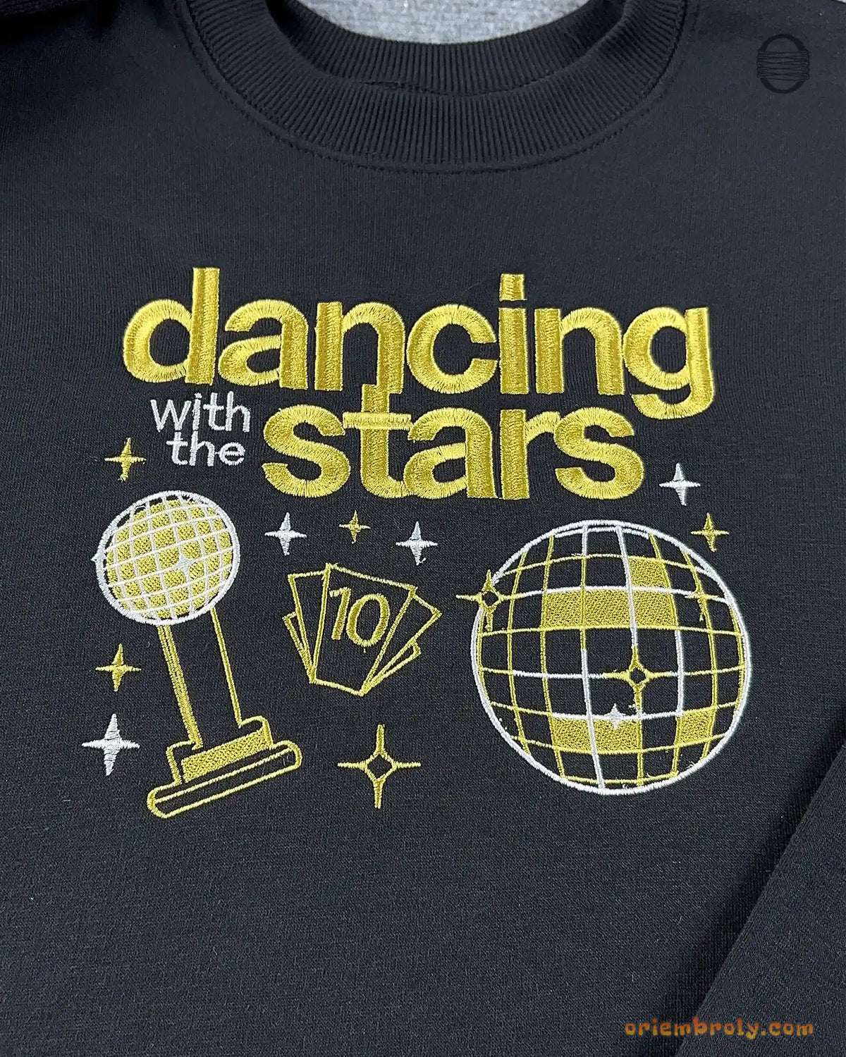 Close-up of gold Dancing with the Stars embroidery featuring mirrorball trophy, dance shoes, and disco ball on a black sweater
