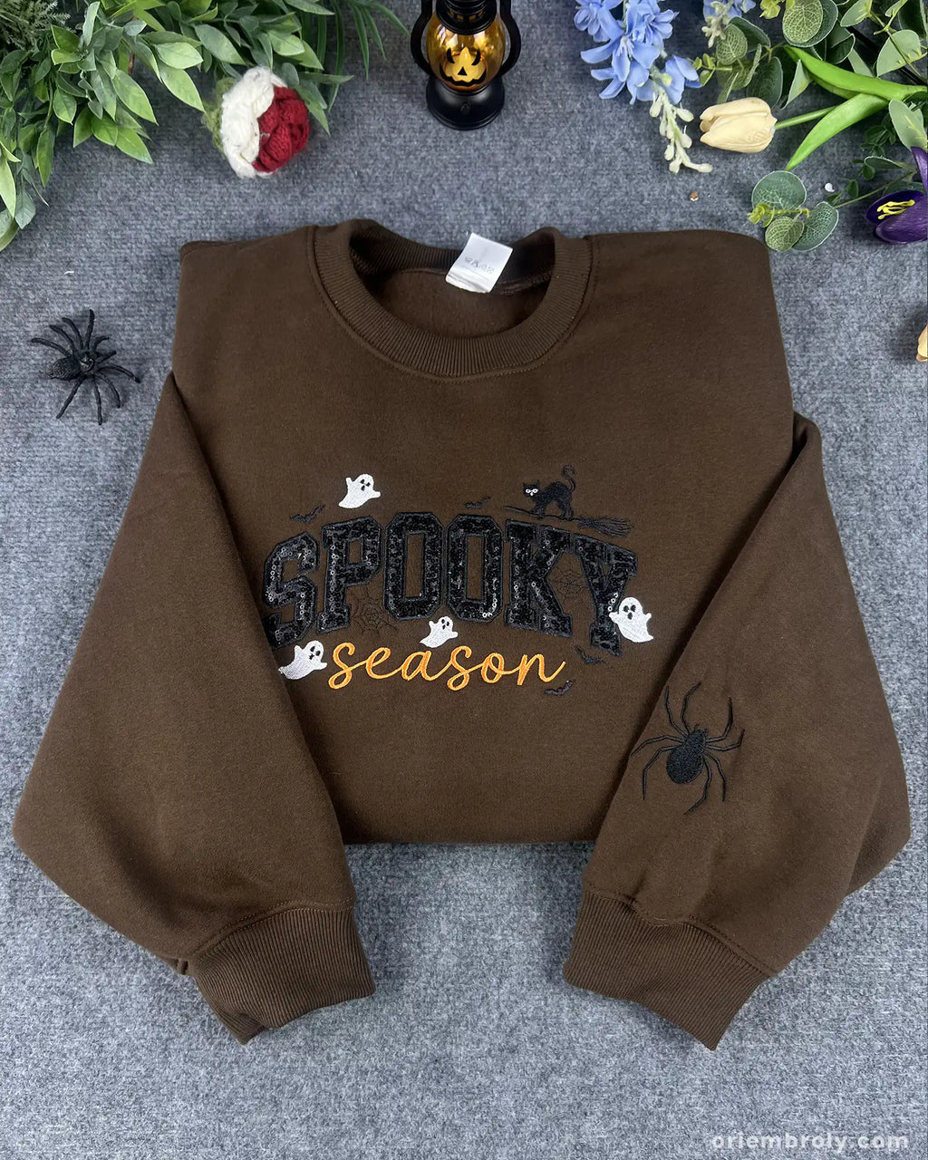 Dark brown spooky season embroidered crewneck sweatshirt flatlay with sequin letters and embroidered spider sleeve.