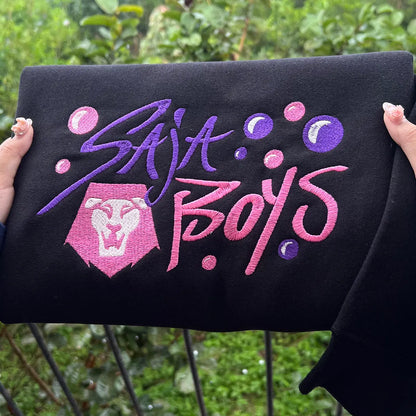 Black sweatshirt with 'Saja Boys' text and logo held by a person outdoors.