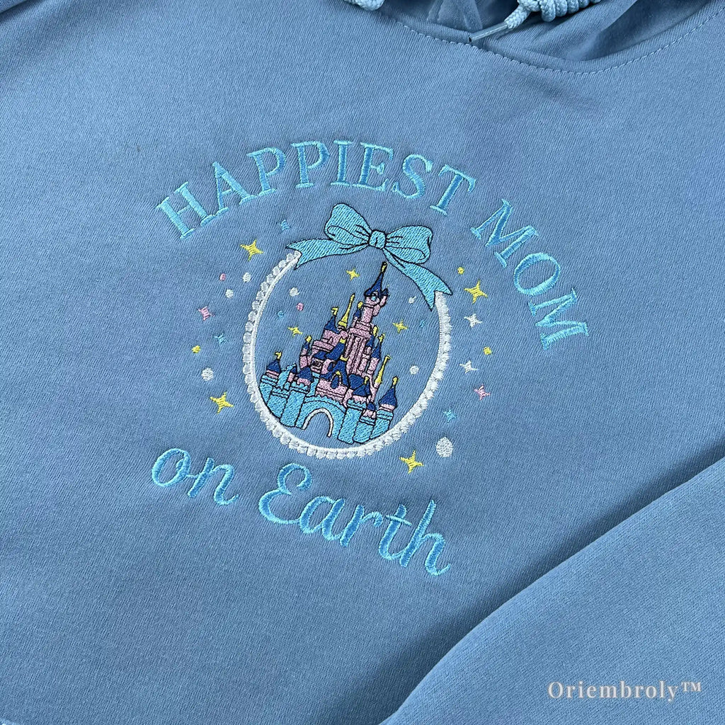 Close-up of premium satin stitch embroidery showing a pink and purple fairytale castle within a pearl-like circular frame on a soft blue hoodie. Durable, high-density thread work