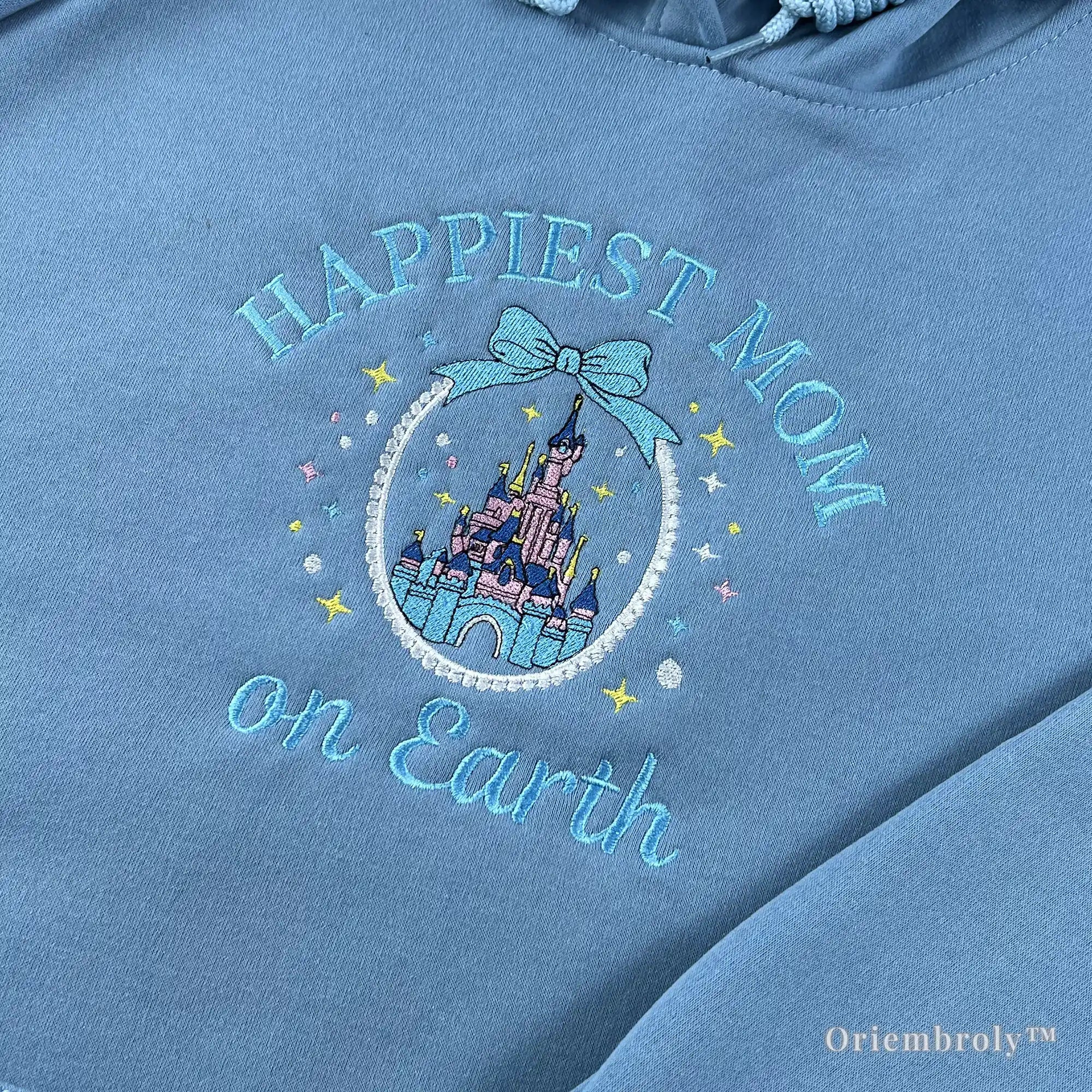 Close-up of premium satin stitch embroidery showing a pink and purple fairytale castle within a pearl-like circular frame on a soft blue hoodie. Durable, high-density thread work