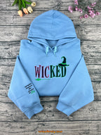 Light blue hoodie with 'WICKED' text on a wooden surface with flowers.