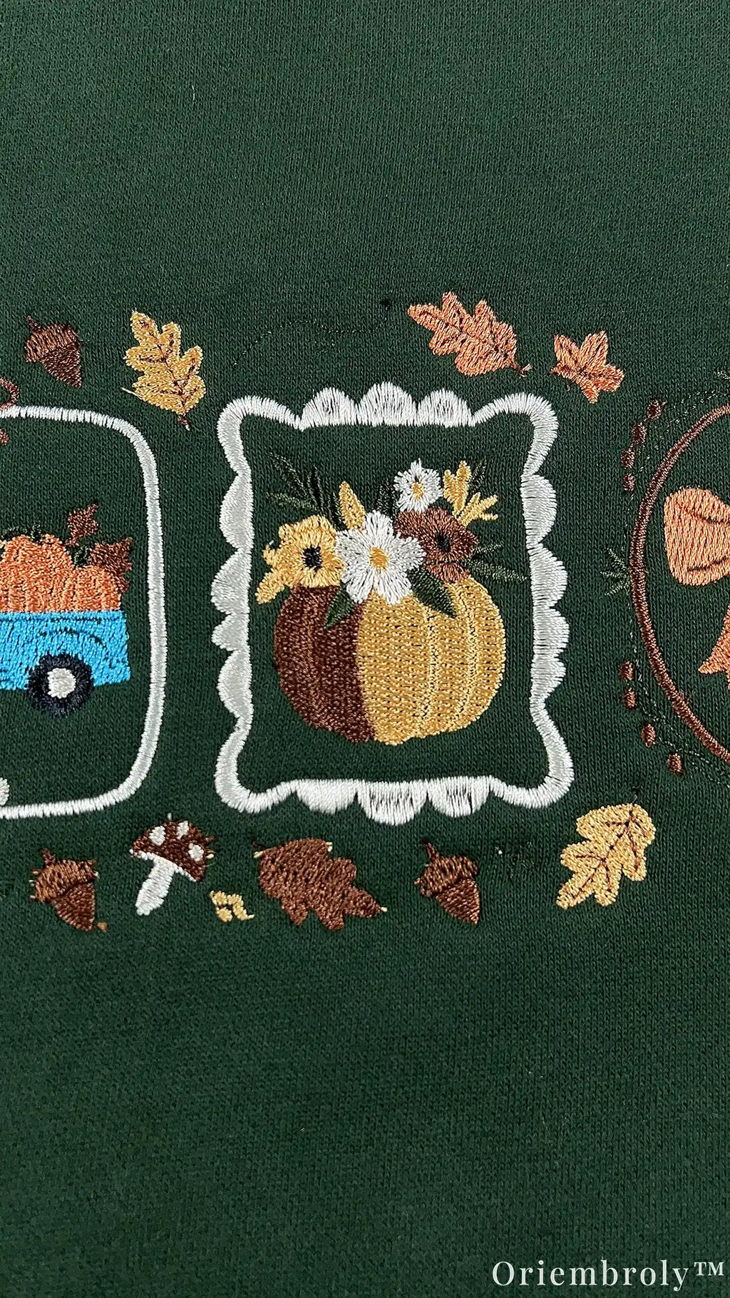 Close-up of embroidered fall pumpkin arrangement inside a white frame surrounded by autumn leaves and tiny mushrooms on green fabric.