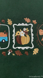 Close-up of embroidered fall pumpkin arrangement inside a white frame surrounded by autumn leaves and tiny mushrooms on green fabric.