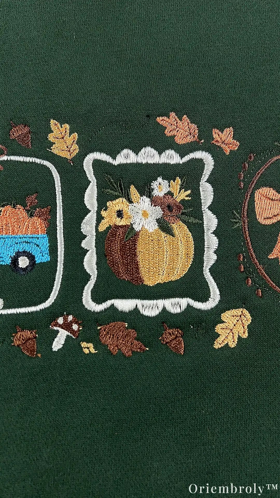 Close-up of embroidered fall pumpkin arrangement inside a white frame surrounded by autumn leaves and tiny mushrooms on green fabric.