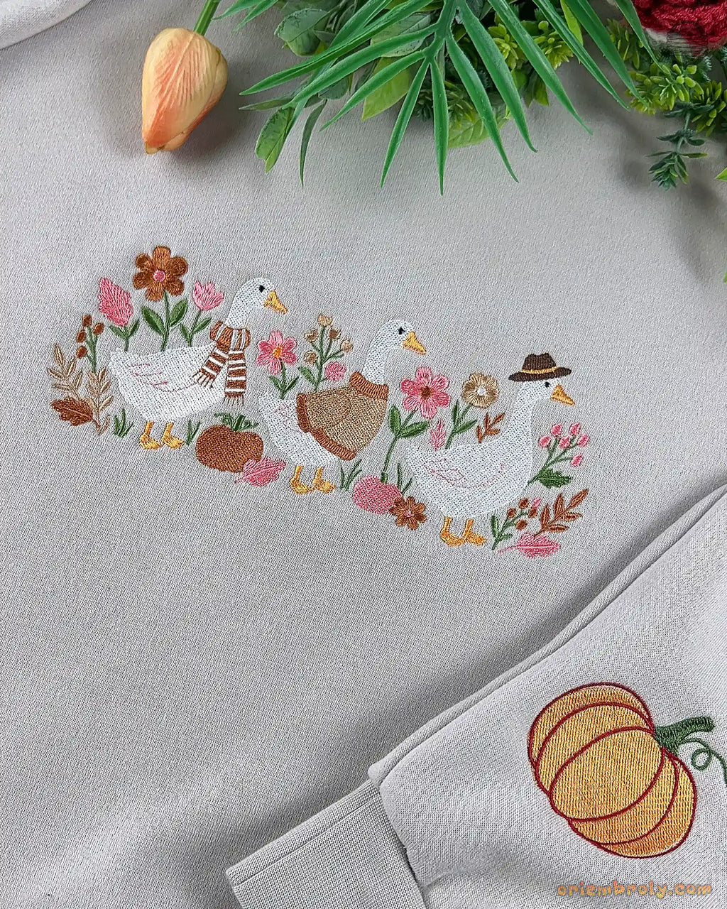 Close-up of fine embroidery showing white ducks wearing fall accessories among flowers, alongside an embroidered orange pumpkin on the sleeve cuff.