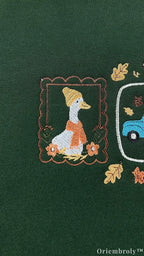 Close-up of fine embroidery showing a framed duck wearing an orange scarf and hat with autumn leaves on a green sweatshirt.